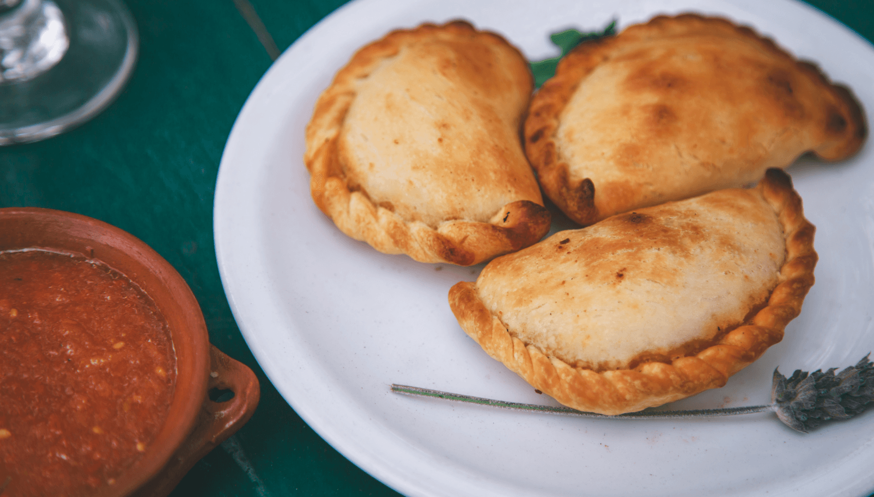 Three baked empanadas on a white plate beside a clay bowl of salsa criolla