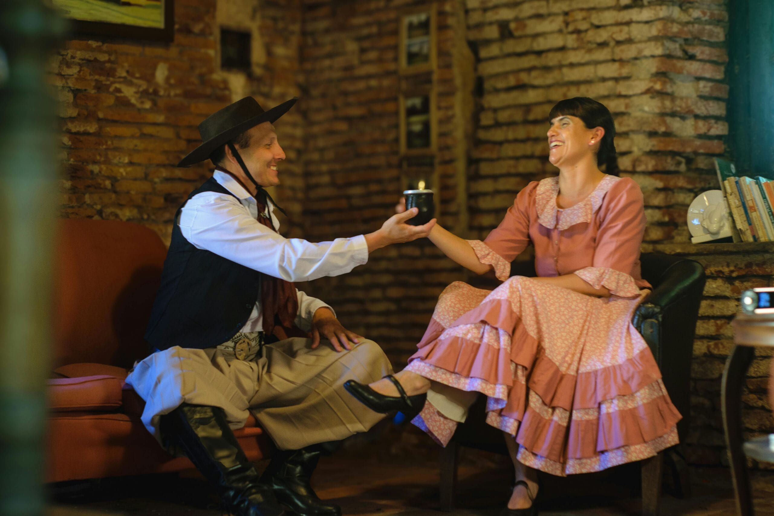 A gaucho in traditional dress passing a mate gourd to a woman in a brick-walled room