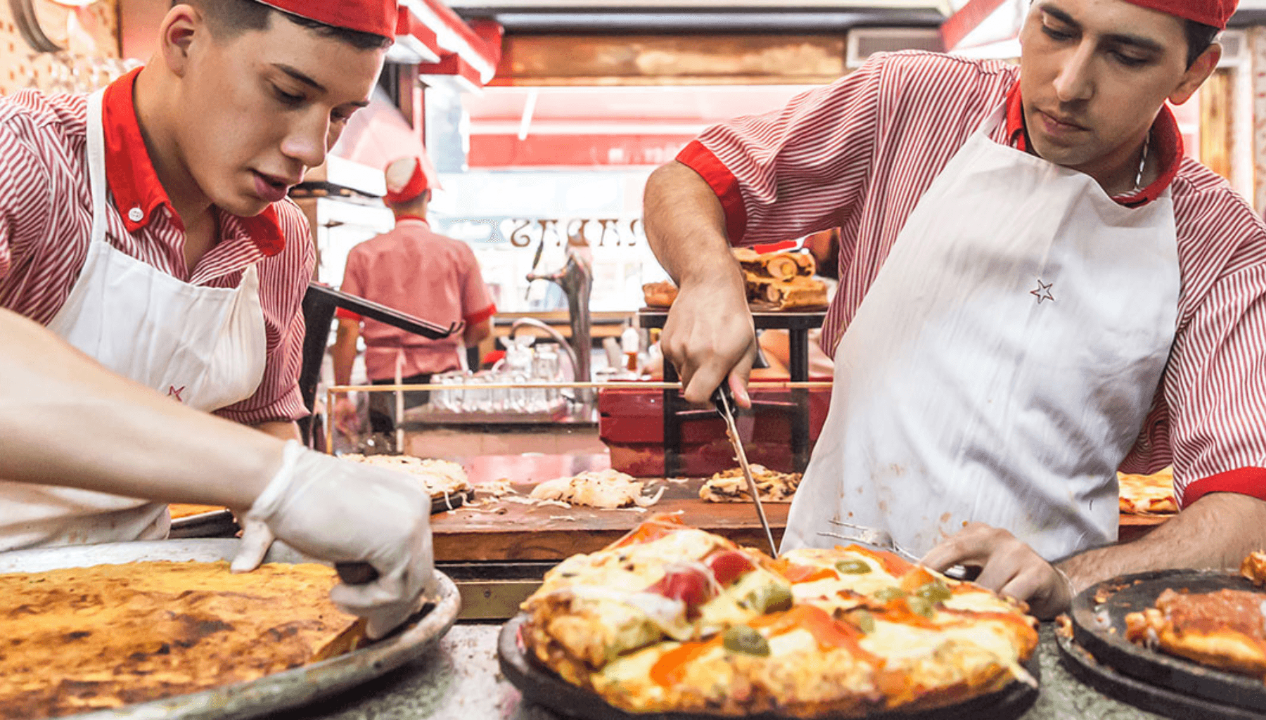 Deux employés de Güerrín en tenue rayée rouge et blanche découpant de la fainá et une pizza garnie au comptoir