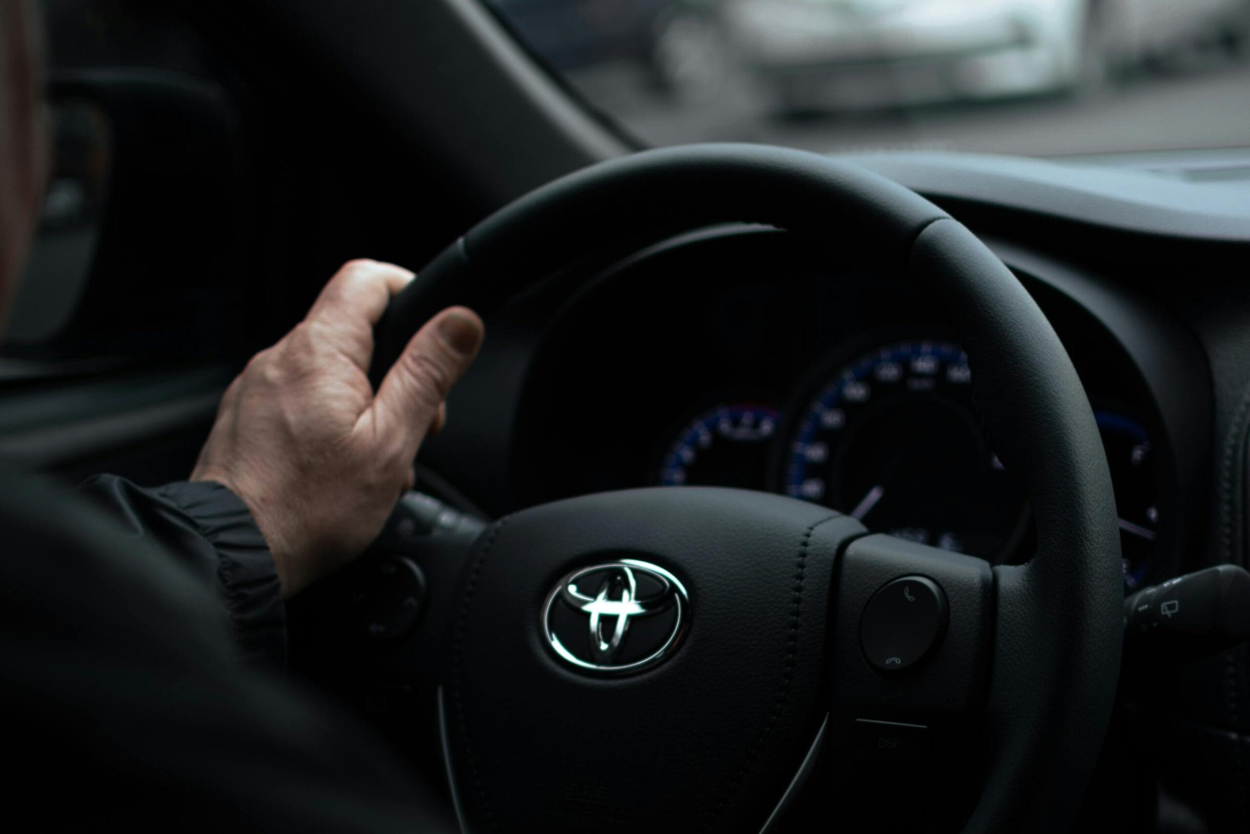 A driver's hand on the steering wheel of a car, with a street and cars blurred through the windscreen