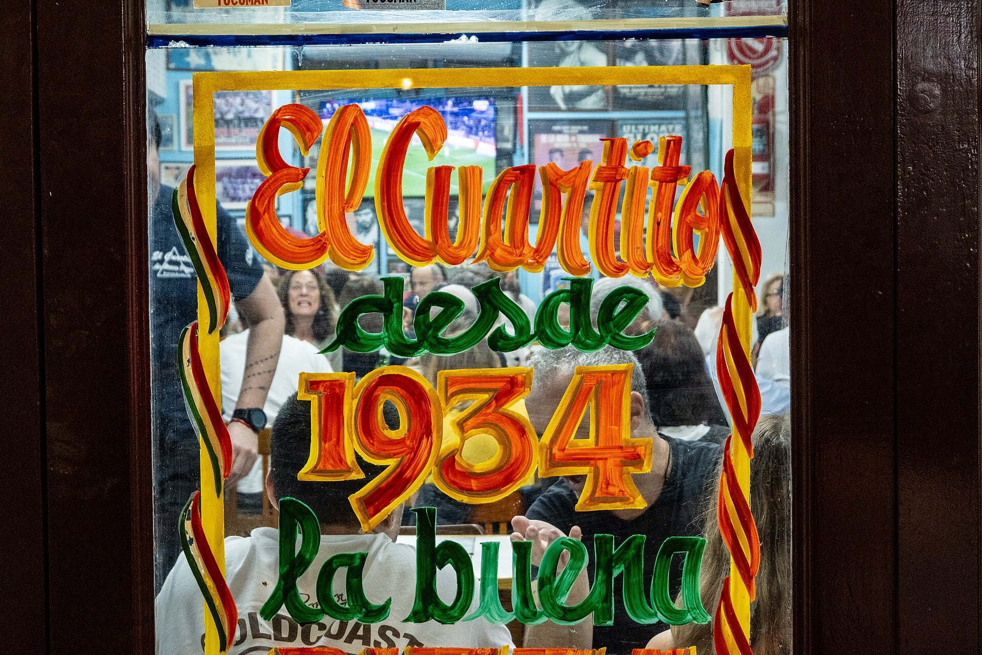A glass door hand-painted in orange, yellow and green with the words El Cuartito desde 1934, and a full dining room with a football match on TV visible through it