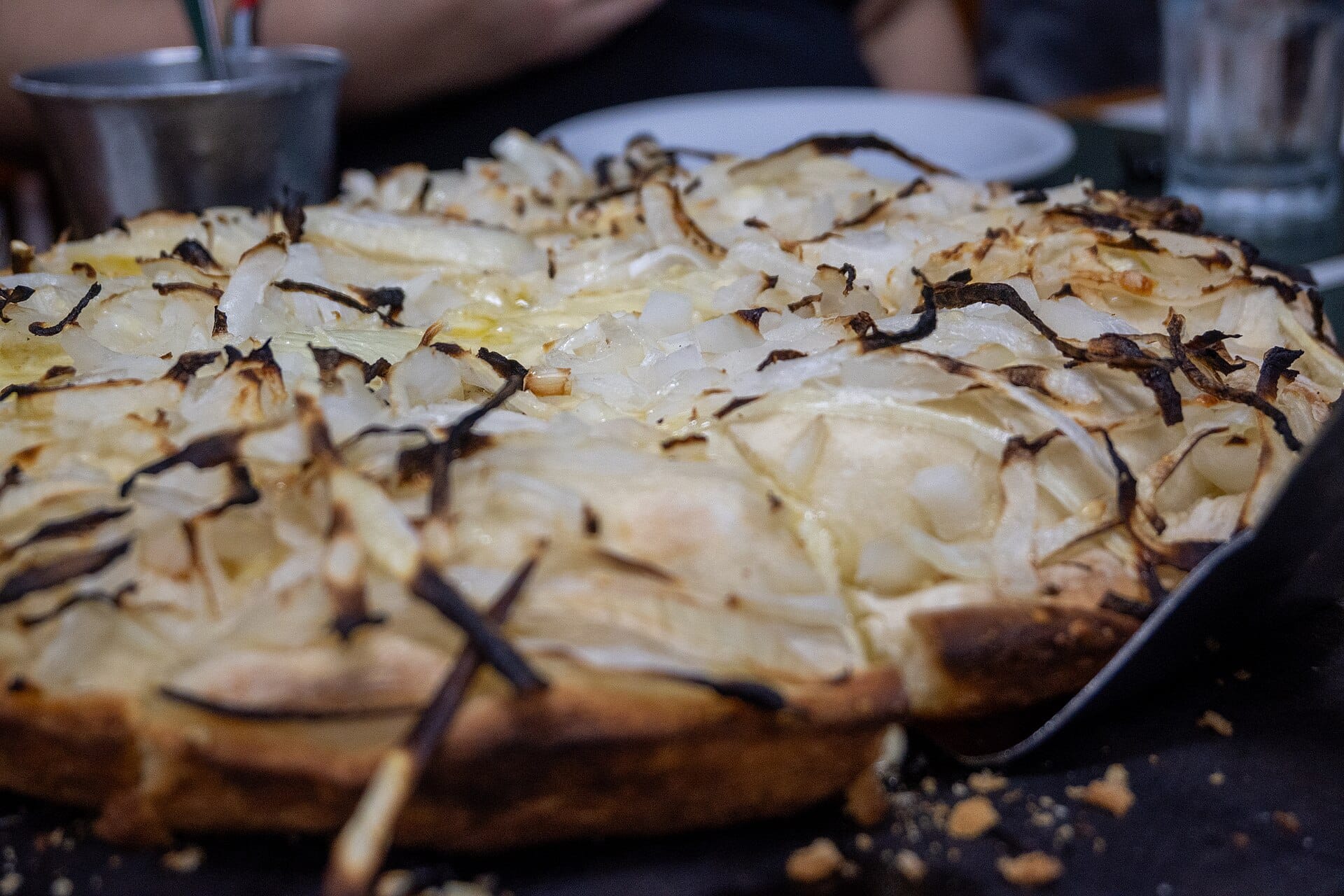 A tall pizza covered in a thick layer of soft onion with charred tips, on a black metal tray, a plate and an aluminium cup behind it