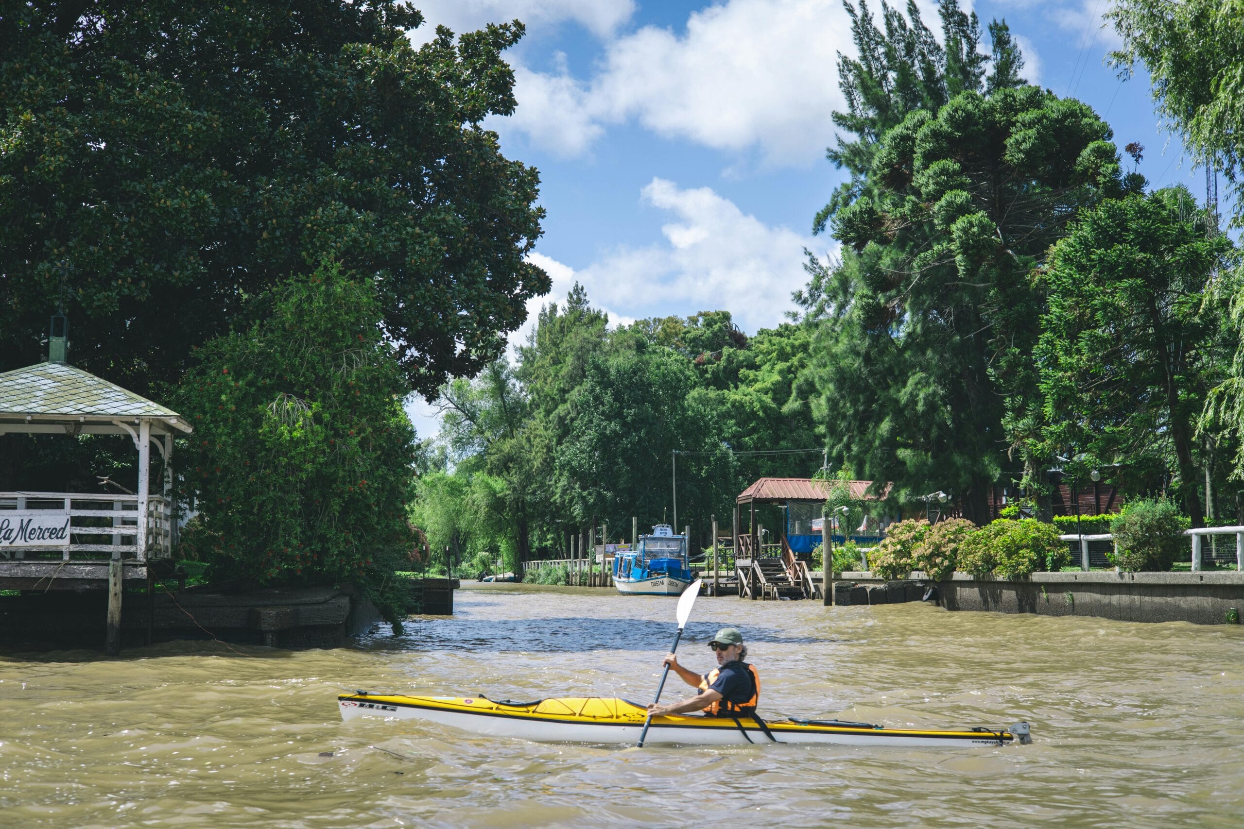 A rower on the Luján river passing a wooden pavilion on the bank