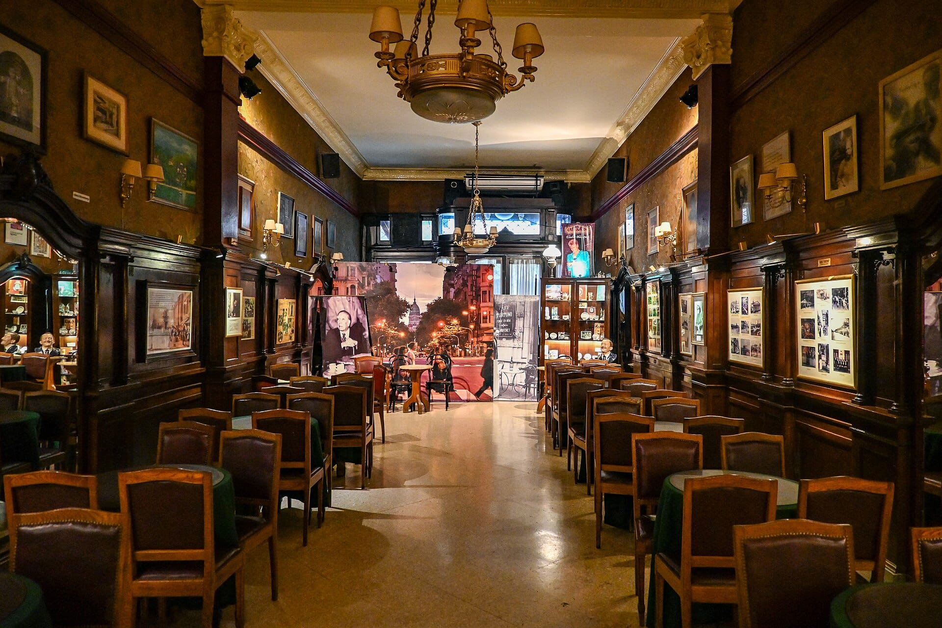 A long wood-panelled café room with rows of empty leather chairs around small green-topped tables, framed pictures on the walls and a brass chandelier overhead