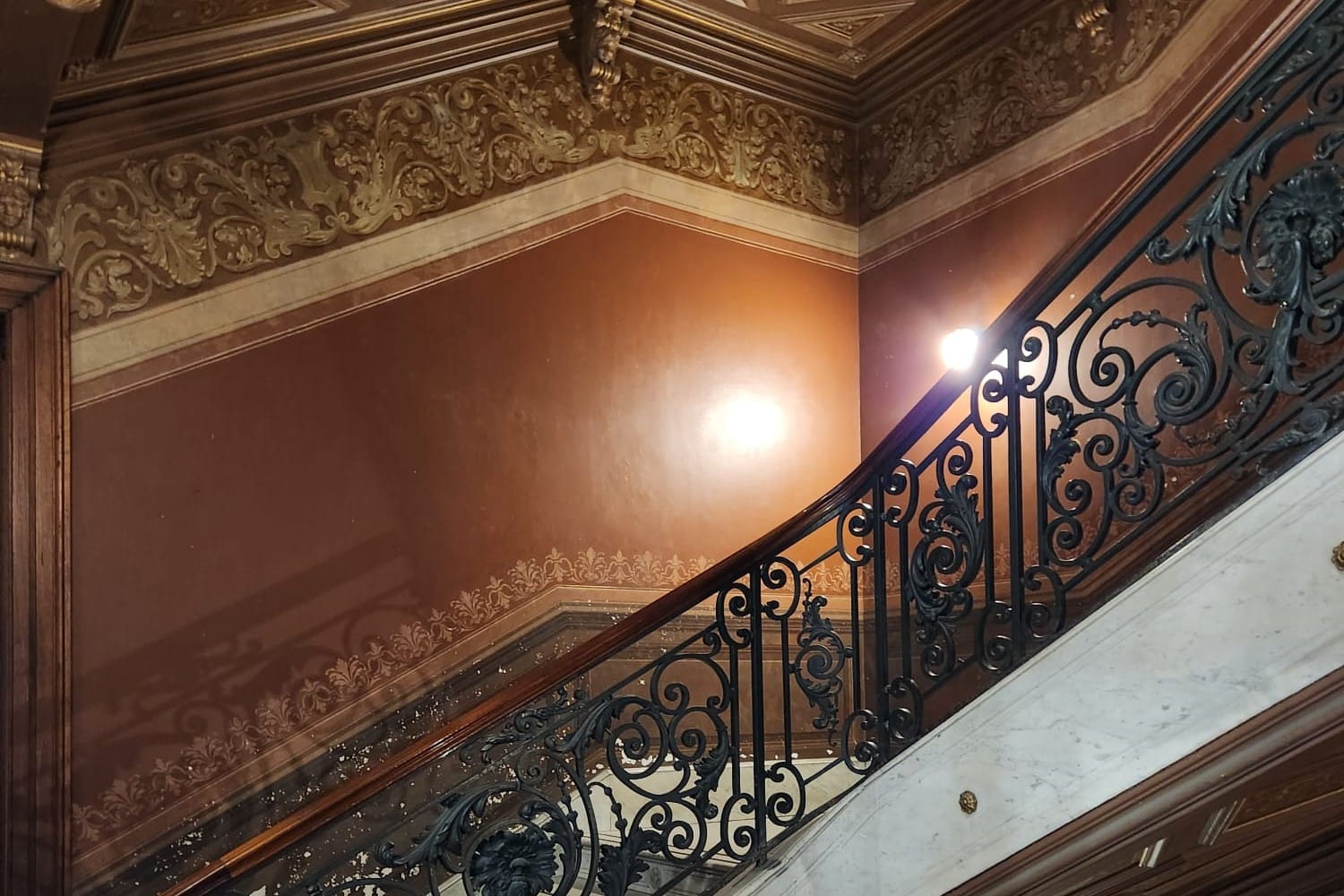 A curving staircase with a wrought-iron railing and a wooden handrail, against warm terracotta walls with gilded friezes
