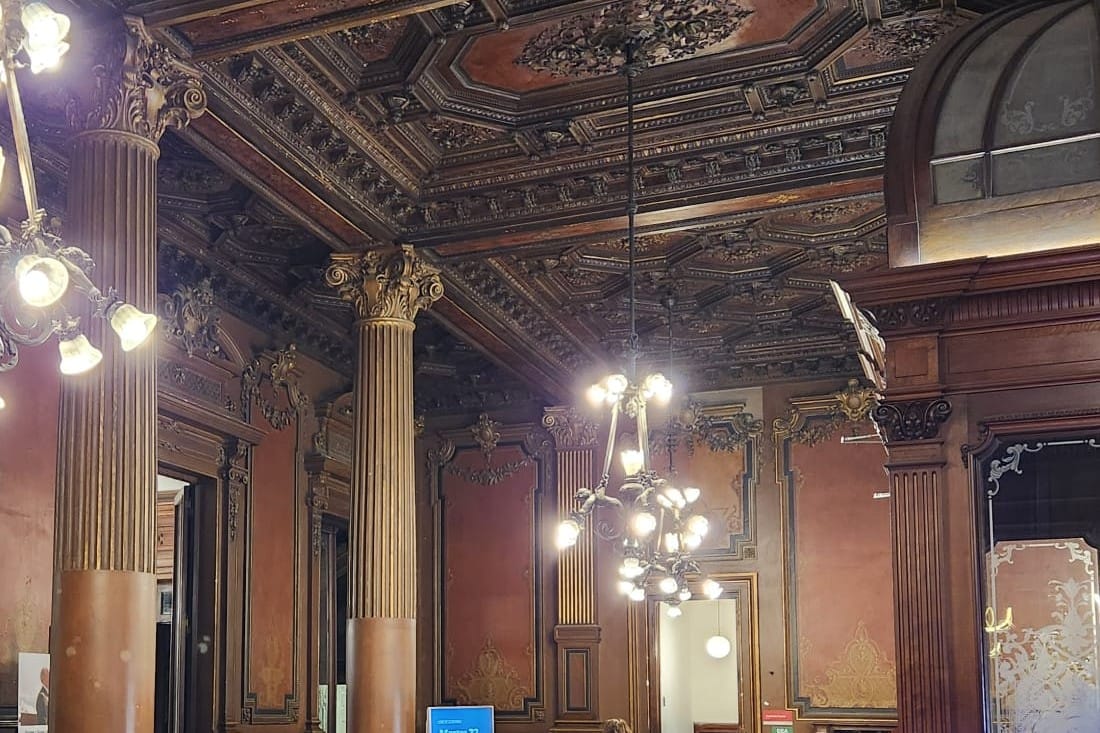 A high ceiling of carved dark wood above a hall with tall columns, wooden doors and two lit chandeliers