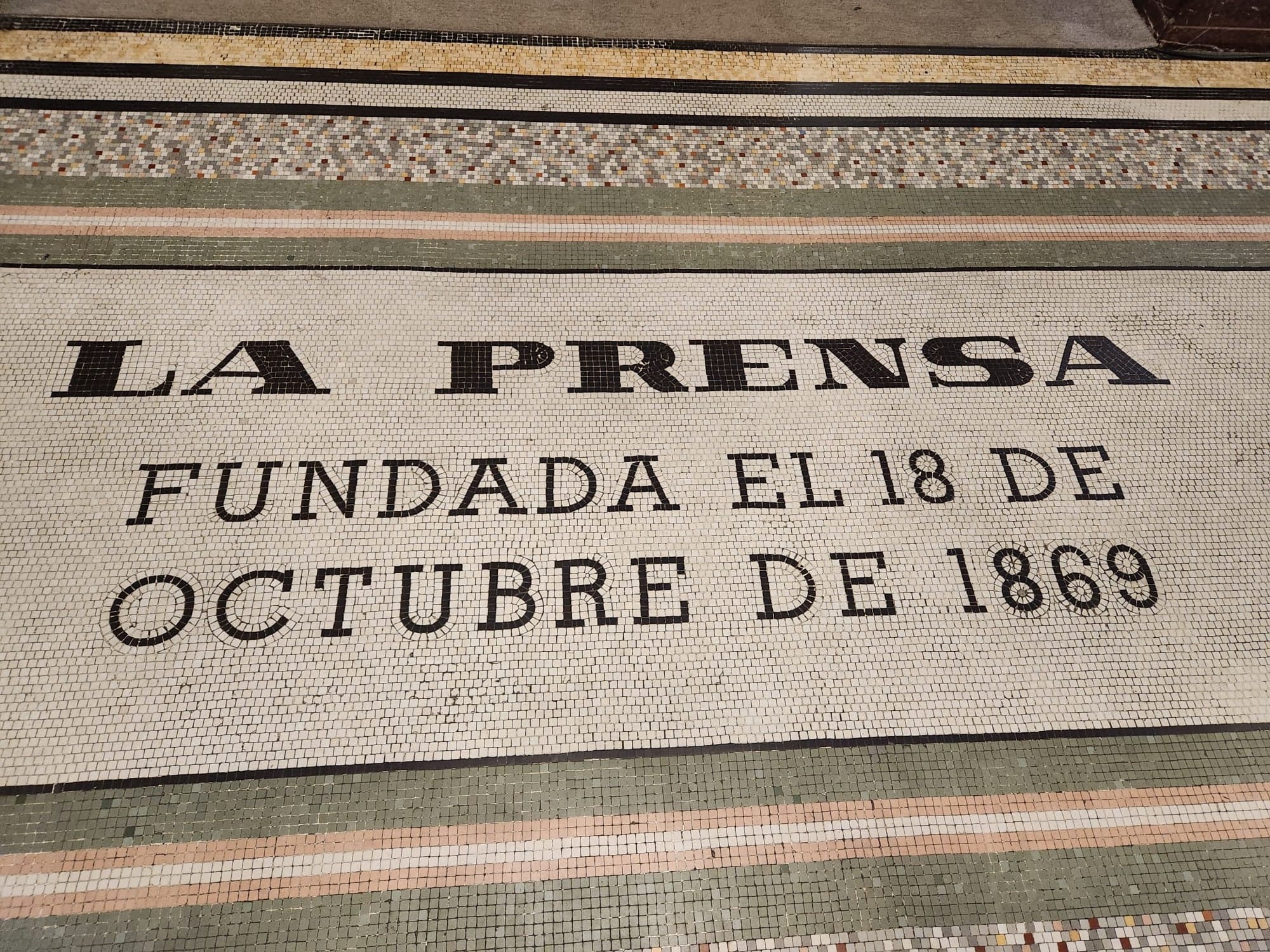 A floor mosaic of small white tiles with the words La Prensa fundada el 18 de octubre de 1869 framed by green and pink bands
