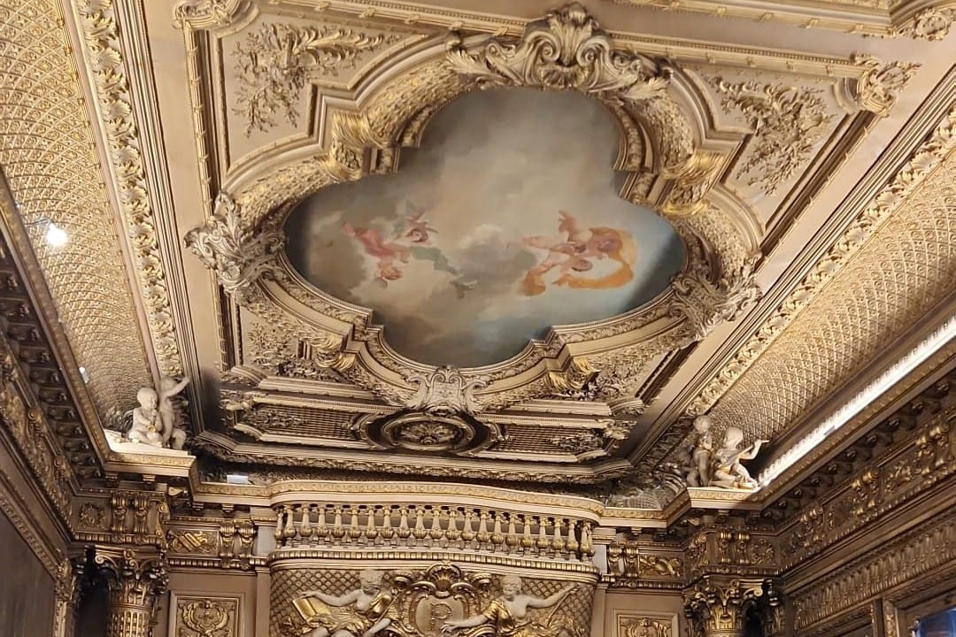 A gilded ceiling with an ornate frame around a painted sky with figures, above gilded walls and small sculpted cherubs