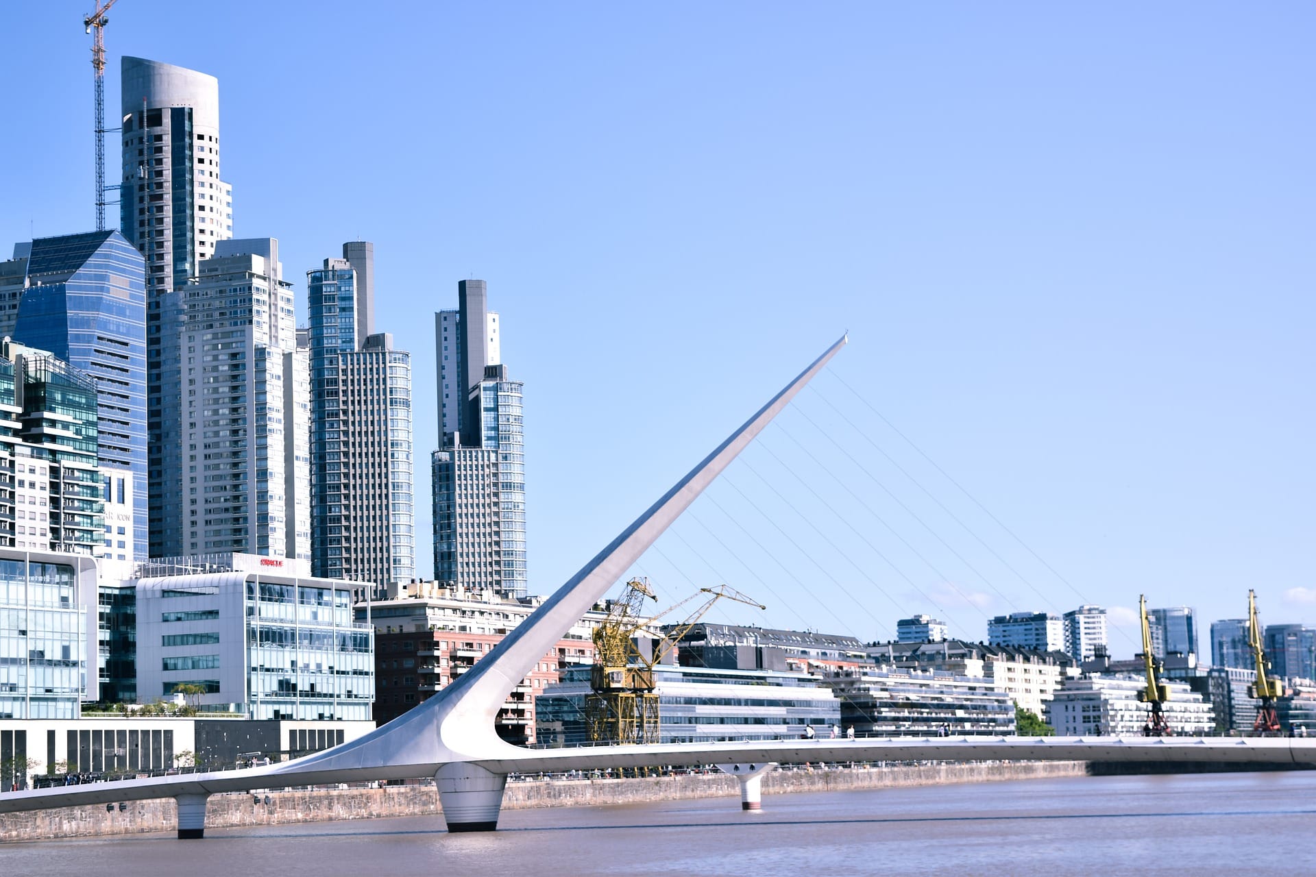 A white footbridge over the docks of Puerto Madero, with glass towers behind it and an orange port crane on the right