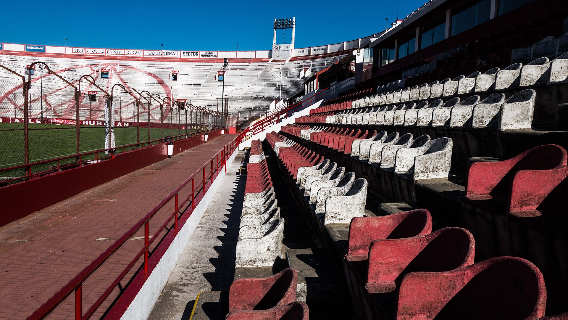 Empty red and white seats in the stands of the Estadio Tomás Adolfo Ducó, with the pitch and a floodlight tower behind them under a clear blue sky