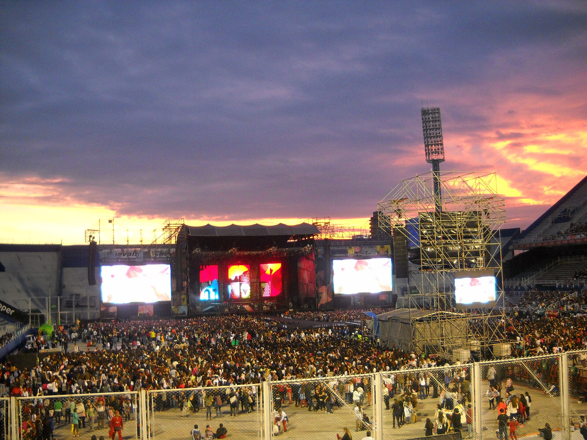 A crowd fills the pitch at Vélez in front of a concert stage with giant screens, under a pink and purple sunset sky