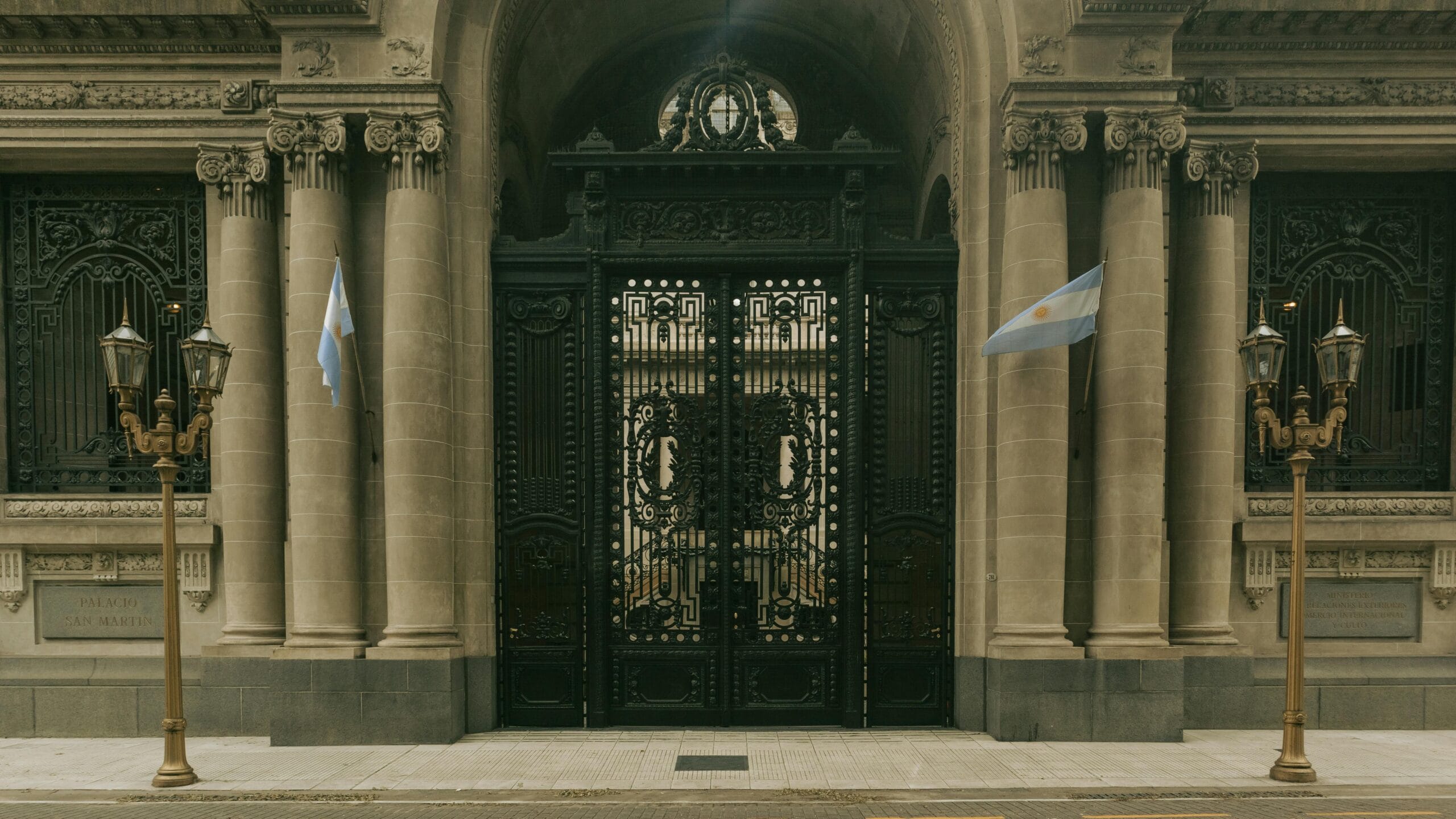 A wrought iron gate in a stone facade with columns, flanked by two Argentine flags and two street lamps