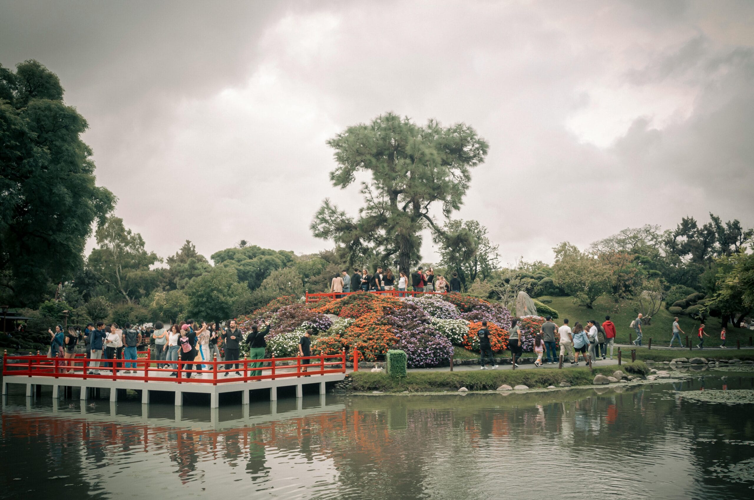 A red wooden bridge and deck over a pond, with visitors on the bridge, a mound of orange and purple flowers and a tall pine under a grey sky